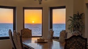 A dining area with a view of the sunset over the sea, a table with chairs, and a potted plant.