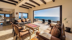 A spacious living area with a large window overlooking the ocean, furnished with leather sofas, wooden chairs, and a coffee table.