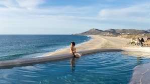 A woman sitting by a pool with a beach and mountains in the background.