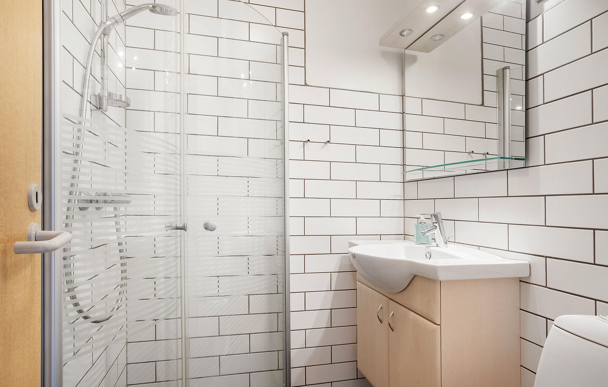 Un baño moderno con azulejos blancos tipo metro, un box de ducha de vidrio, un tocador color beige con un lavabo y un espejo sobre el lavabo.