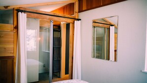 Un dormitorio con cama, armario de madera, puerta corredera de vidrio que da a un baño y un espejo en la pared.