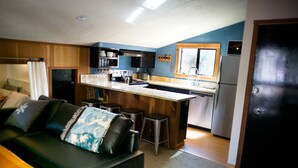 Un espacio habitable compacto con kitchenette, un sofá con cojines decorativos y una mesa de comedor de madera.