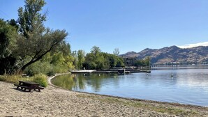 A sandy beach with a picnic table and a dock extending into a calm lake, surrounded by trees and hills in the background. - Luxury top-floor Penthouse with private dock, pool and stunning lake views. (Osoyoos)