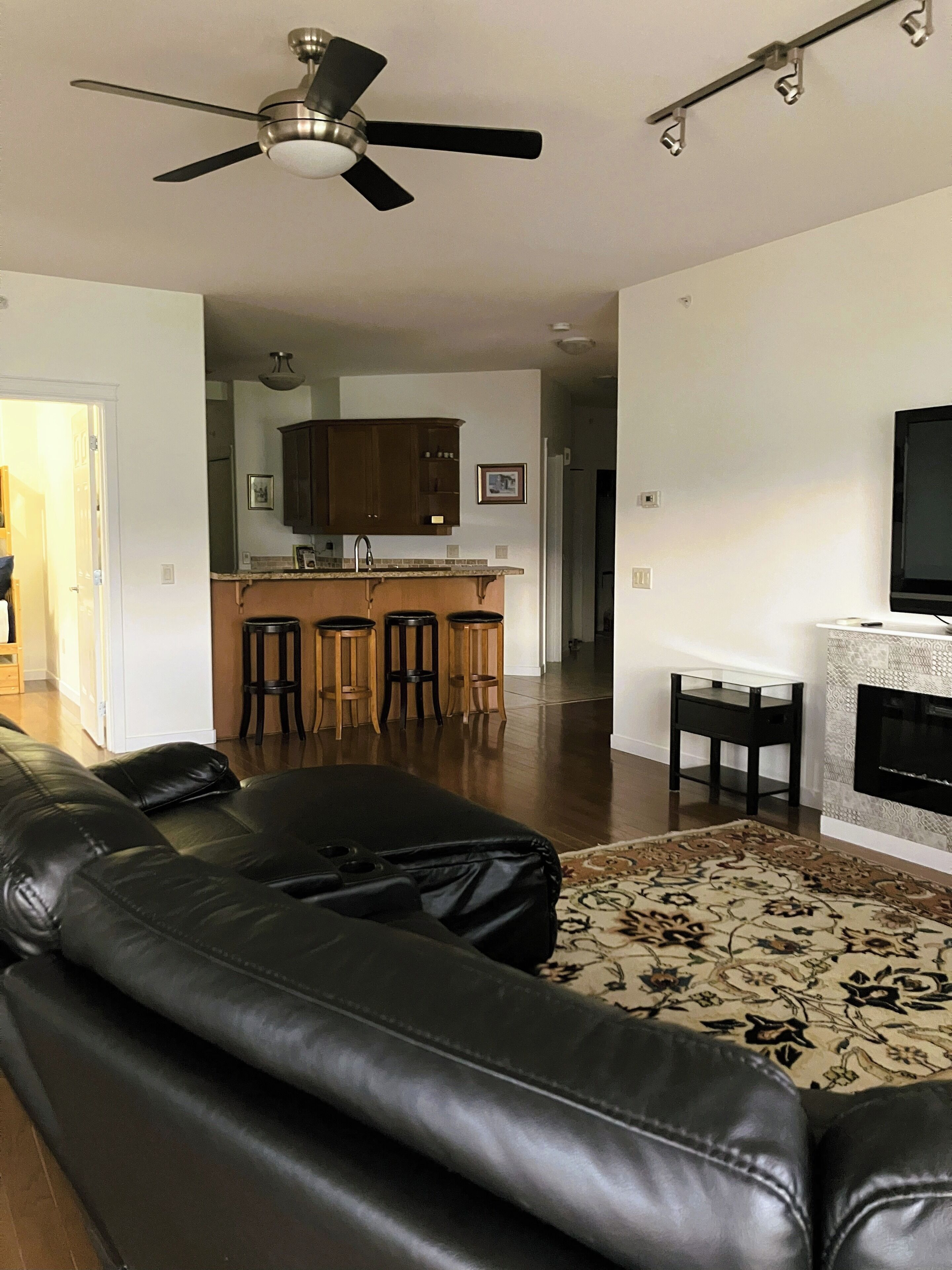 A living room with a black leather sofa, a ceiling fan, a kitchen area with wooden cabinets, a bar stool, and a fireplace.
