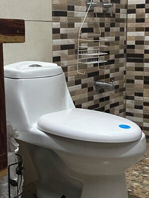 Sanitário com caixa de água e assento brancos, prateleira de metal acima e parede revestida com padrão de mosaico.