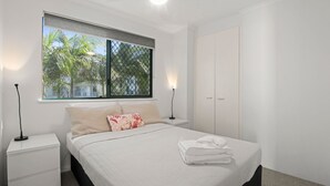 Un dormitorio con cama, mesitas de noche, una ventana con vista a palmeras y un armario.