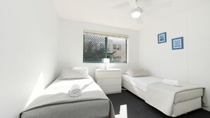 A small, bright room with two single beds, a nightstand, a window with a view of buildings, and a ceiling fan. - Lindomare Unit 6, 11 Orvieto Tce, Kings Beach (Kings Beach)