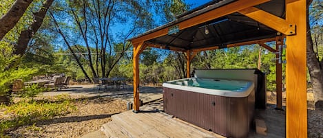 Un jacuzzi bajo una pérgola de madera en un área boscosa con mobiliario exterior.
