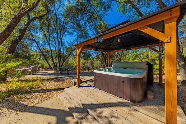 A hot tub under a wooden pergola in a wooded area with outdoor seating.