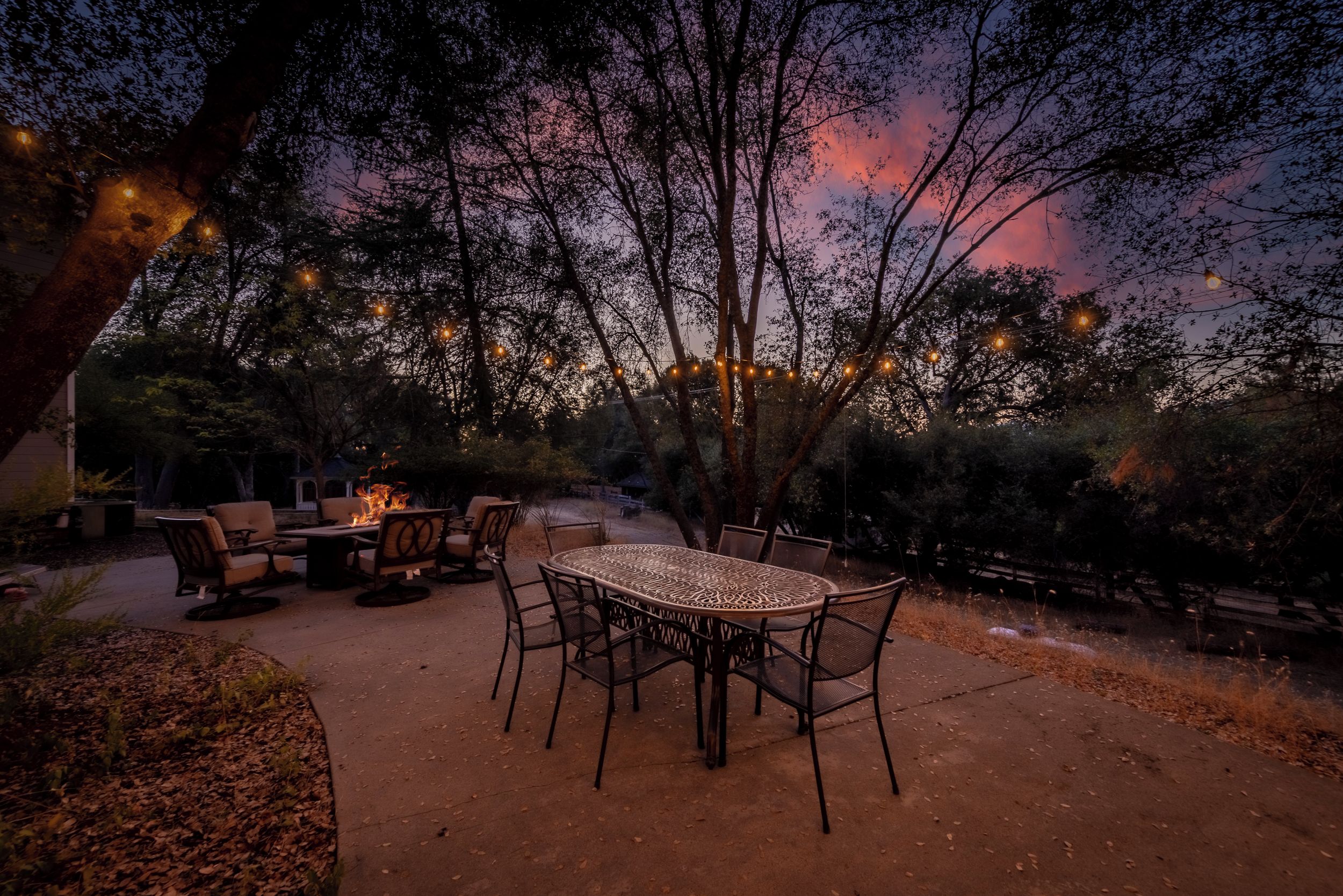An outdoor patio with a round table and chairs, surrounded by trees and string lights.