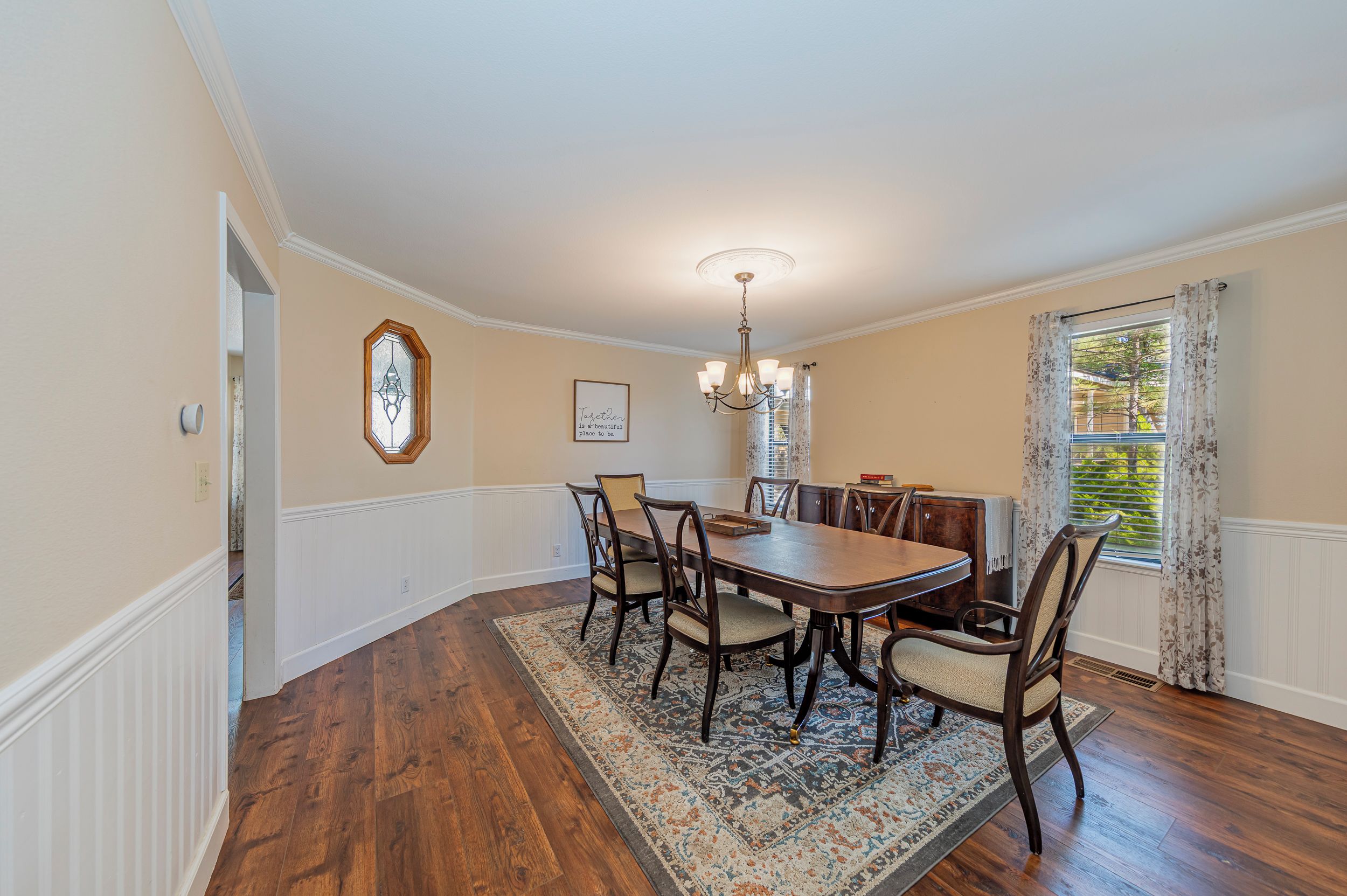 A dining room with a wooden table and chairs, a chandelier, a rug, and a window with curtains.