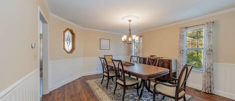 A dining room with a wooden table and chairs, a chandelier, a rug, and a window with curtains.