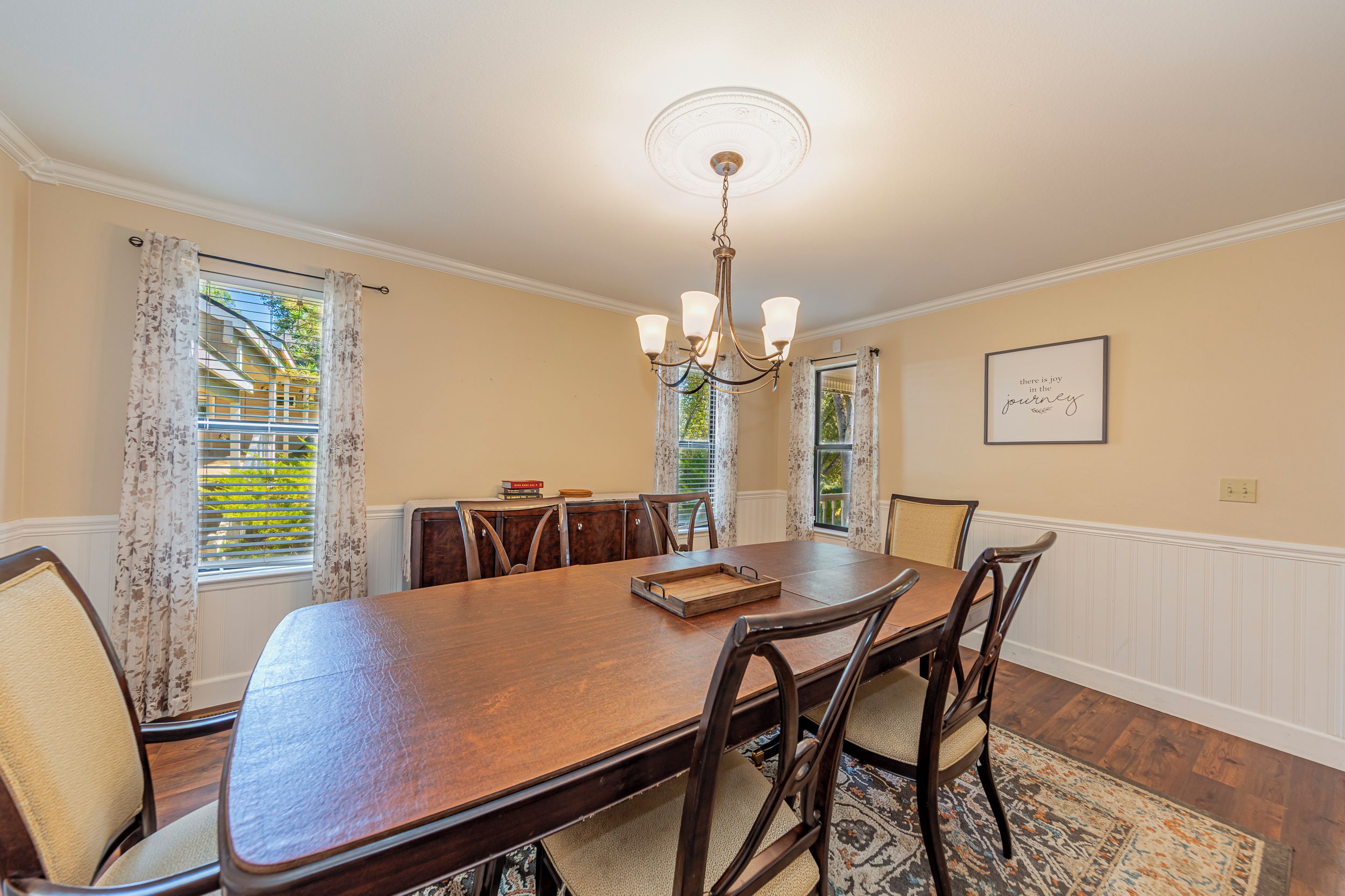 A dining room with a large wooden table, six chairs, a chandelier, and a framed quote on the wall.