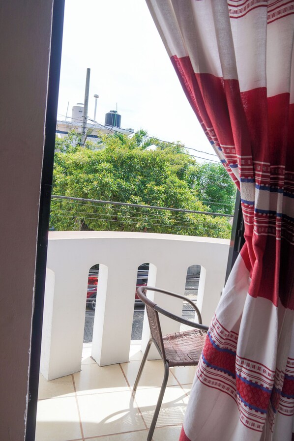 Un balcón con una silla de metal, vistas a zonas verdes y edificios, y una cortina roja y blanca que cubre parcialmente la ventana.