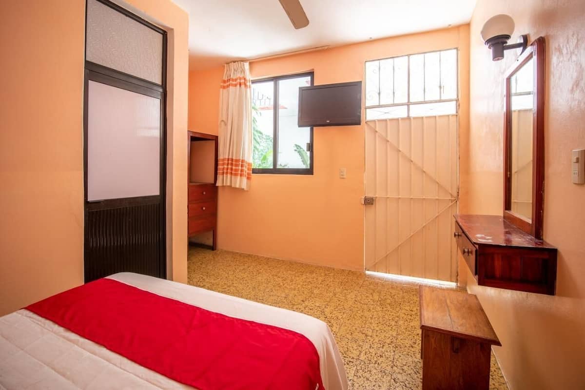 A hotel room with a bed, a wooden bench, a TV mounted on the wall, and a window with curtains.