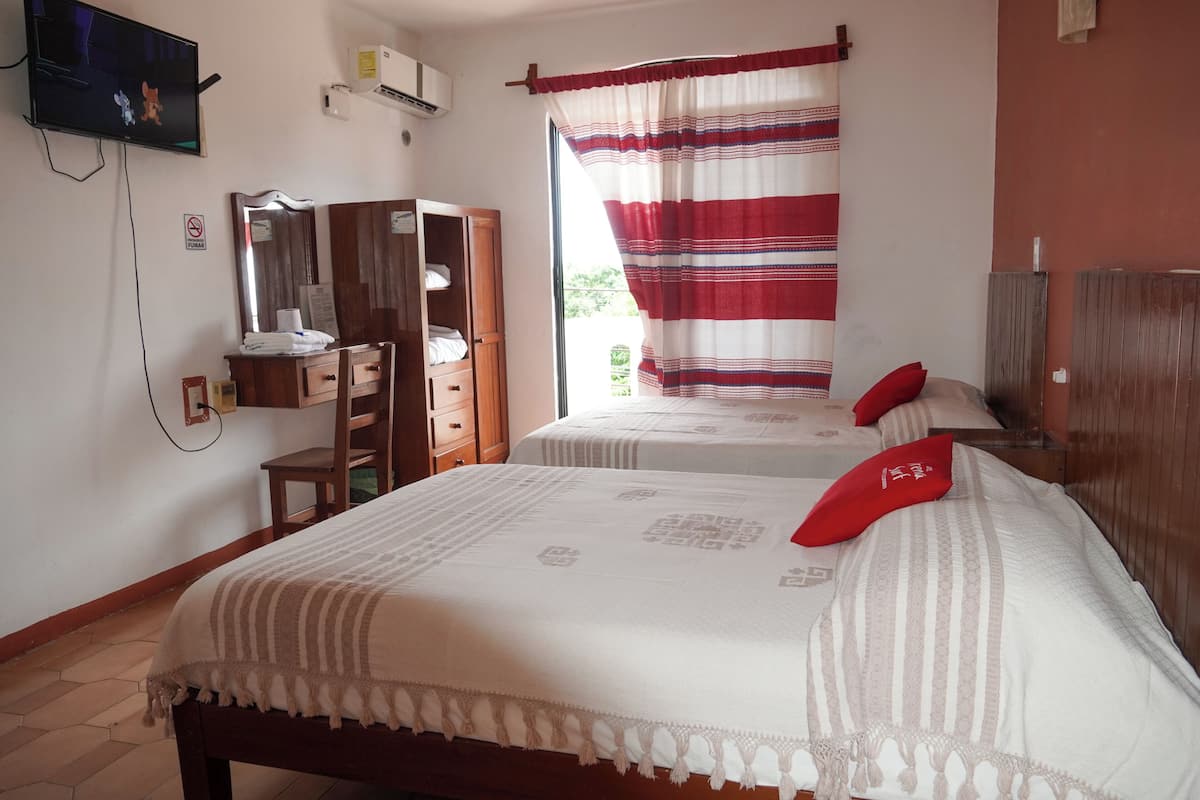 A hotel room with two beds, a wooden wardrobe, a mirror, a television, and a window with striped curtains.