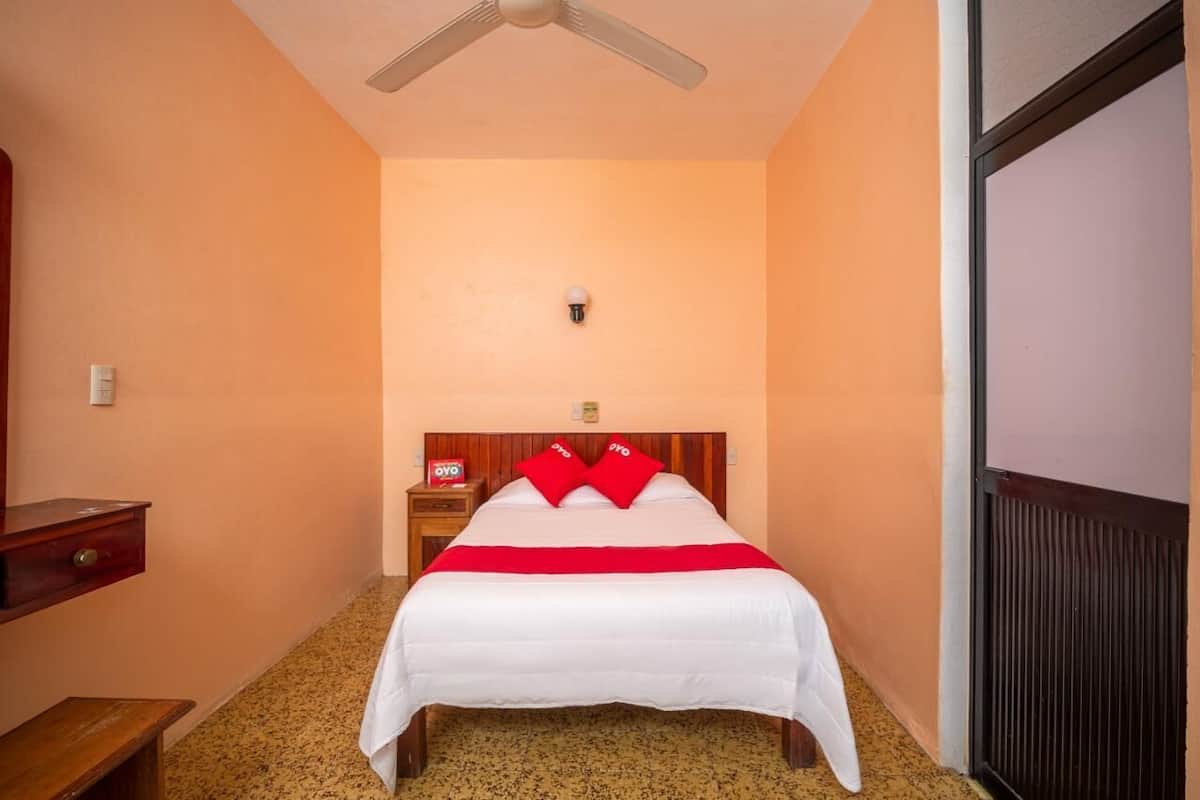 A hotel room with a bed, a nightstand, a wooden bench, a ceiling fan, and a radiator.