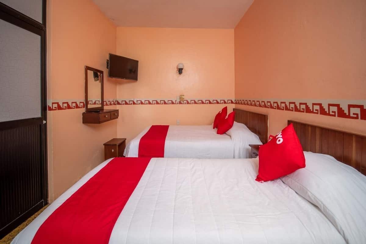 A hotel room with two beds, each with a red and white bedspread, a wooden headboard, a small wooden nightstand, a mirror, and a wall-mounted TV.