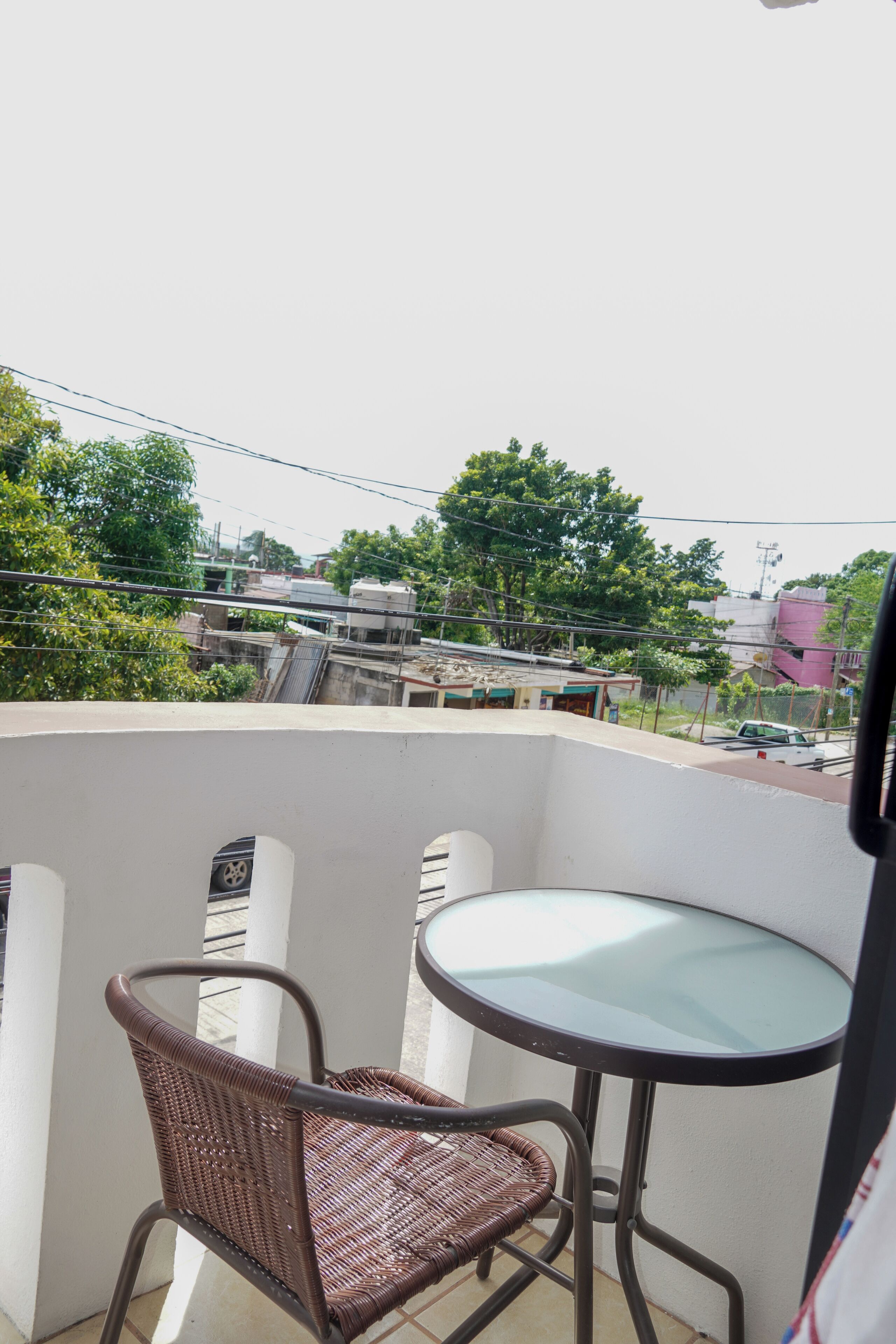 Un balcón con una silla de mimbre y una mesita, con vistas a una zona residencial con árboles y edificios.