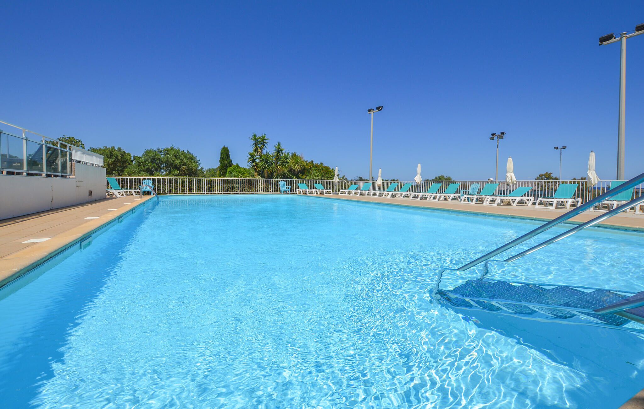 Piscina azul cristalina com cadeiras e guarda-sóis brancos à beira, cercada por um muro branco e palmeiras.