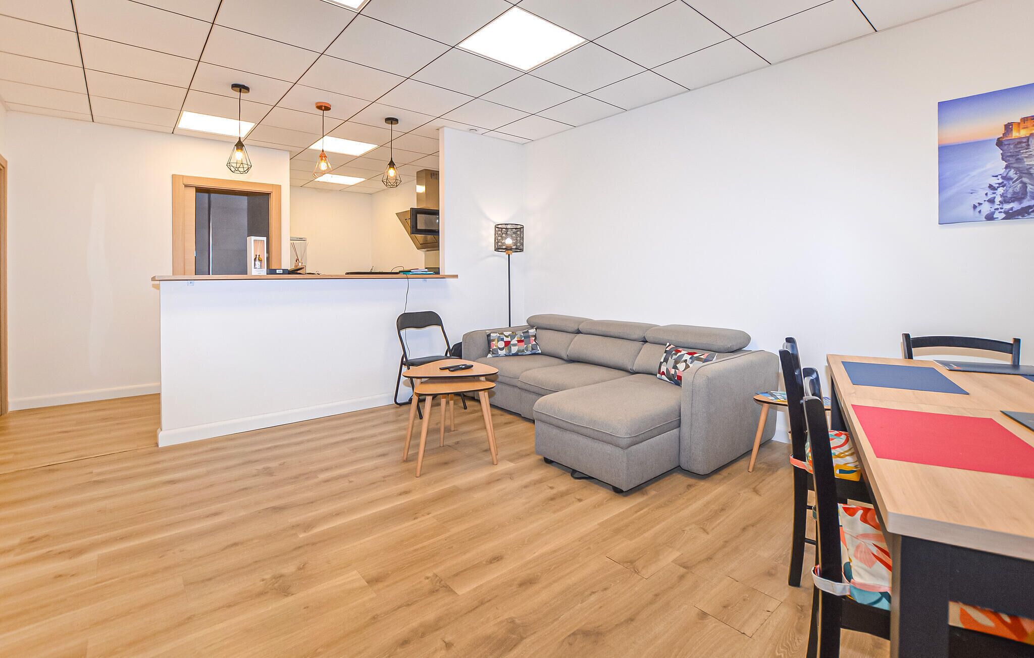 Un salon moderne avec un canapé d’angle gris, une table à manger en bois et un petit coin cuisine équipé d’un réfrigérateur et d’un évier.
