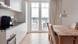 Et moderne køkken med hvide skabe, en sort bordplade og en indbygget ovn. Der er et spiseområde med et træbord og stole, samt et stort vindue med udsigt over byen.