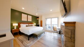 A bedroom with a large bed, bedside tables, a TV mounted on the wall, a stone fireplace, and a balcony with a chair.