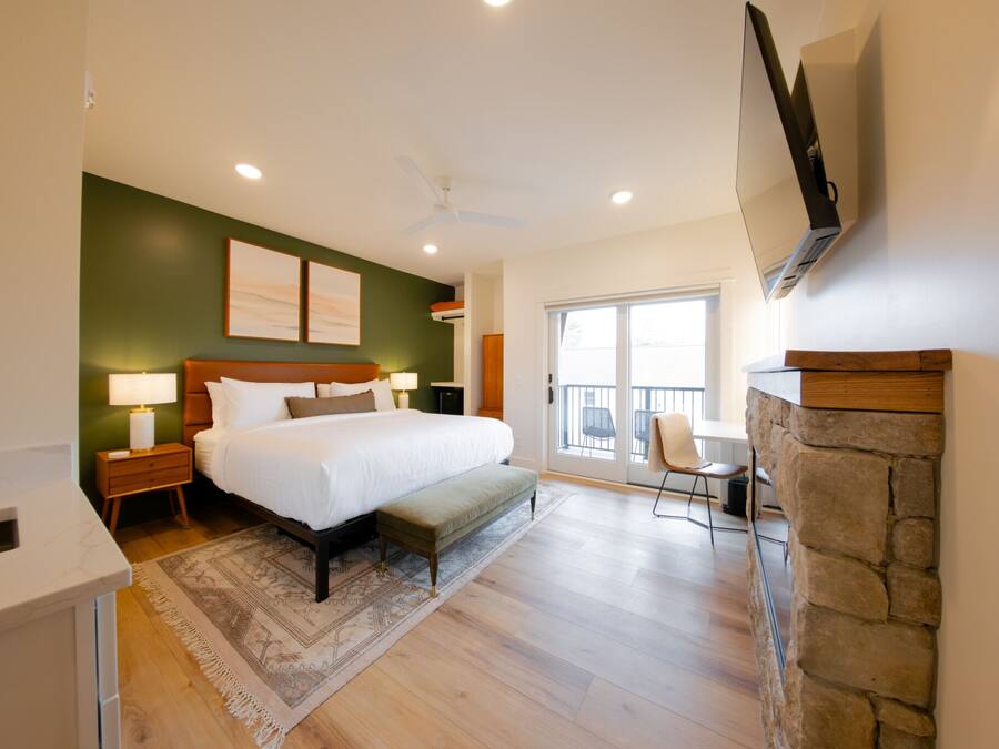A bedroom with a large bed, bedside tables, a TV mounted on the wall, a stone fireplace, and a balcony with a chair.