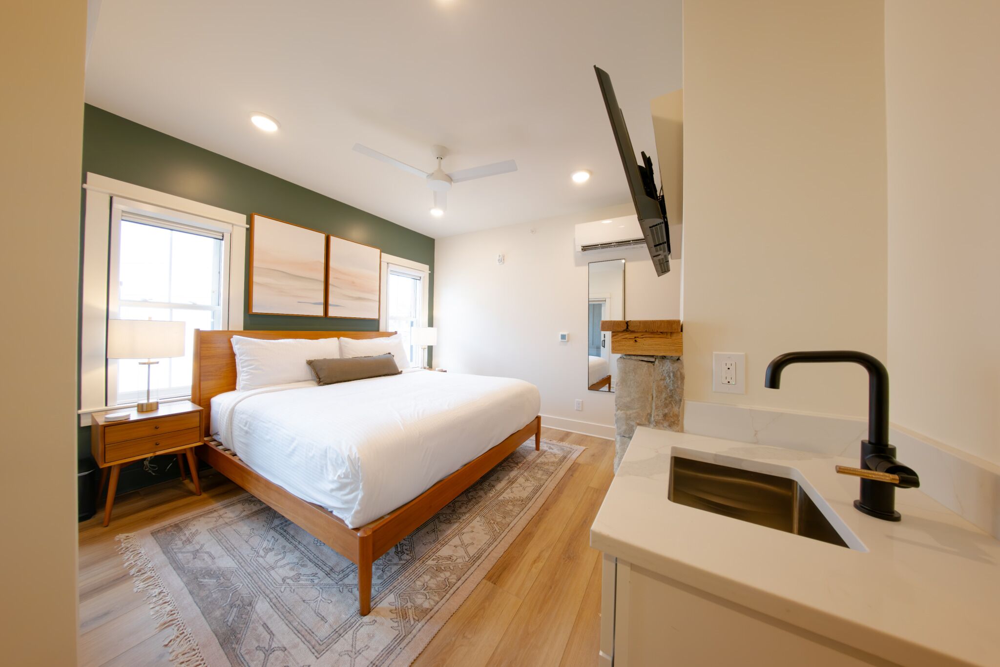 Quarto com cama, mesas de cabeceira, ventilador de teto, janela e banheiro com pia e espelho.