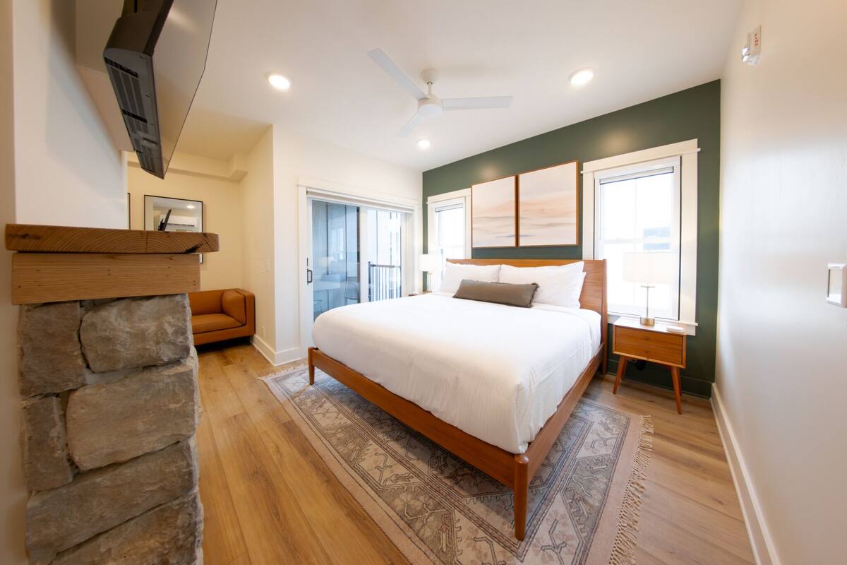 Un dormitorio con cama, mesita de noche, pared de piedra decorativa, banco de madera y vistas al exterior.