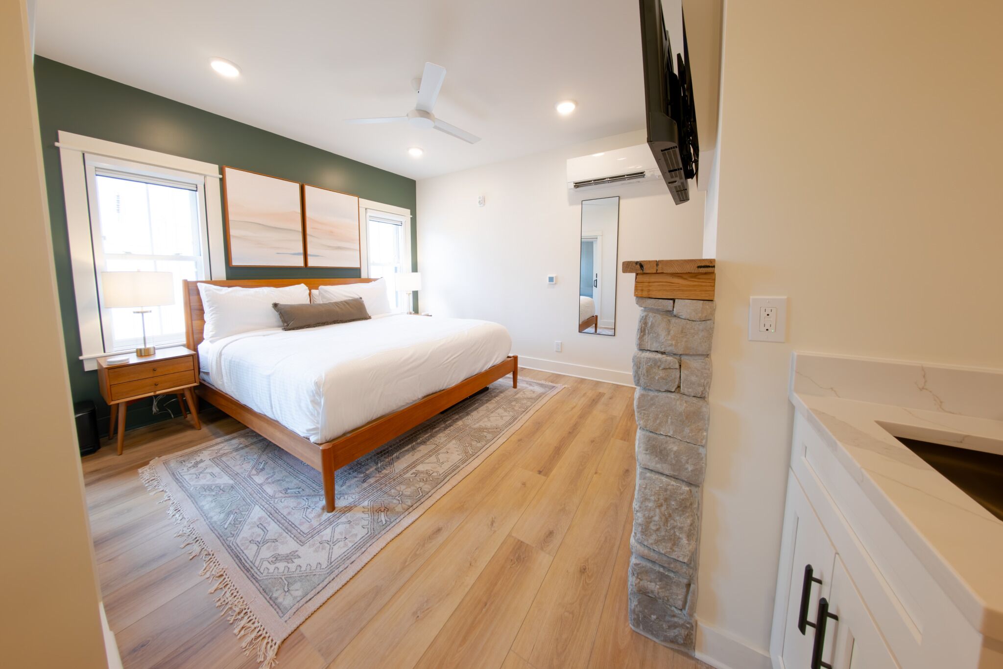 A bedroom with a bed, nightstands, a rug, a stone fireplace, and a large window.
