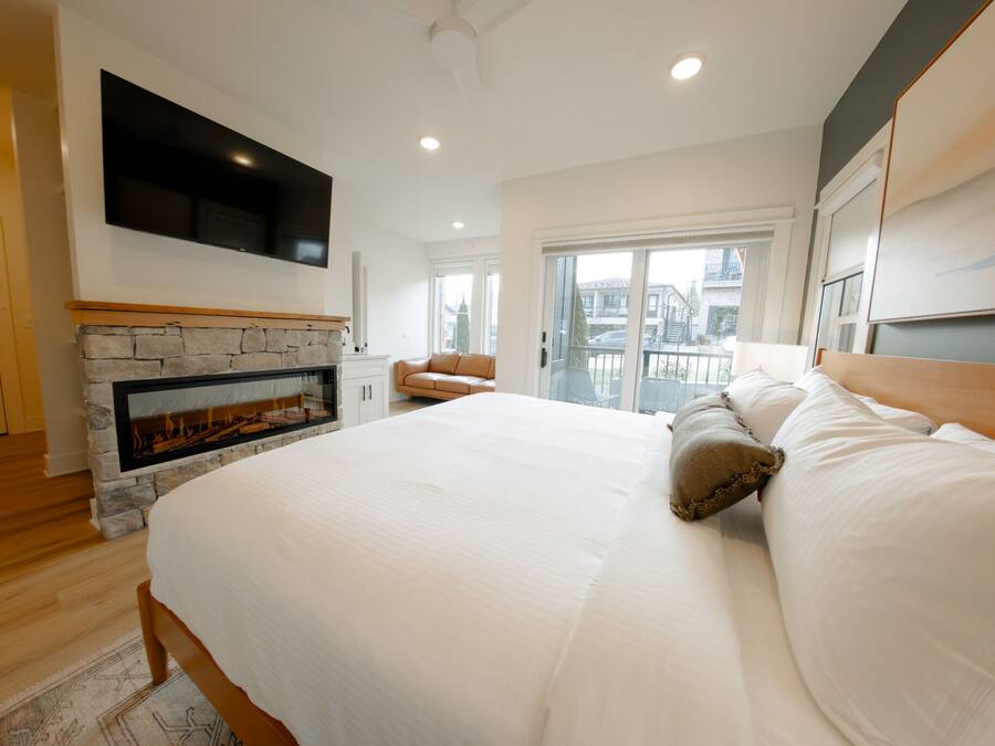 A bedroom with a large bed, a stone fireplace, a TV mounted on the wall, and a balcony with a view of the city.
