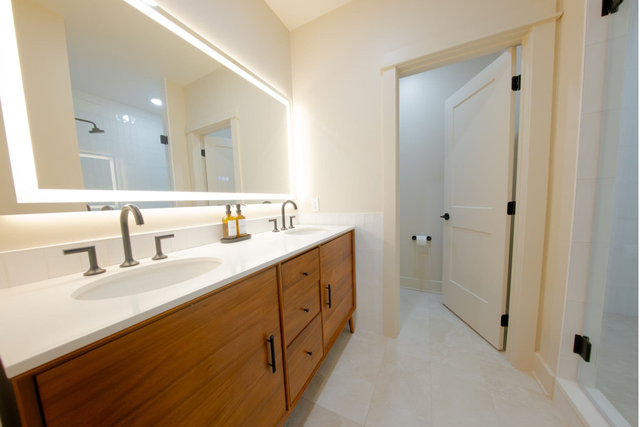 A modern bathroom with a large mirror, two sinks, wooden cabinetry, and a door leading to another room.