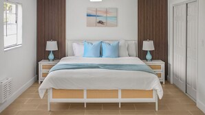 Een slaapkamer met een bed, twee nachtkastjes met lampen, een plafondventilator en een kunstwerk met een strandthema aan de muur.