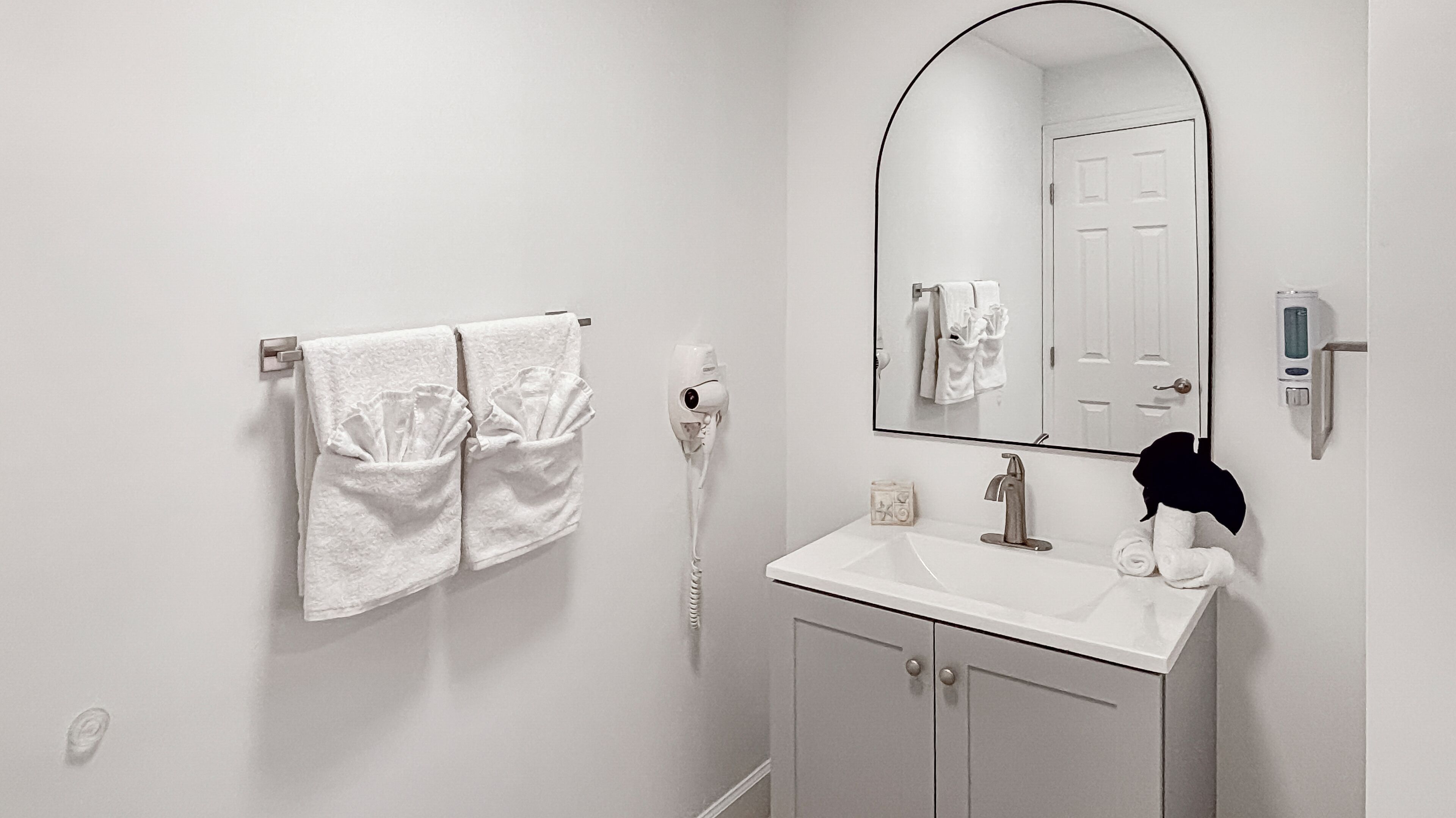 A bathroom with a white sink, a mirror, a towel rack with two towels, a hairdryer, and a soap dispenser.
