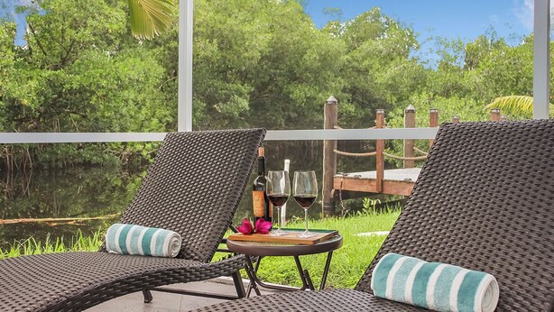 Twee rieten lounge stoelen met gestreepte kussens, een klein rond tafeltje met een boek en twee wijnglazen, en een uitzicht op een waterpartij en groen door grote ramen.