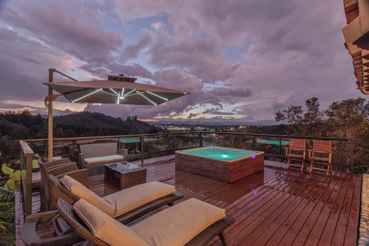 Une terrasse en bois avec des chaises longues, un jacuzzi et une balustrade en verre donnant sur un paysage pittoresque.