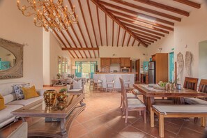 A spacious living area with a high ceiling, wooden beams, a chandelier, and a dining area with a wooden table and chairs.