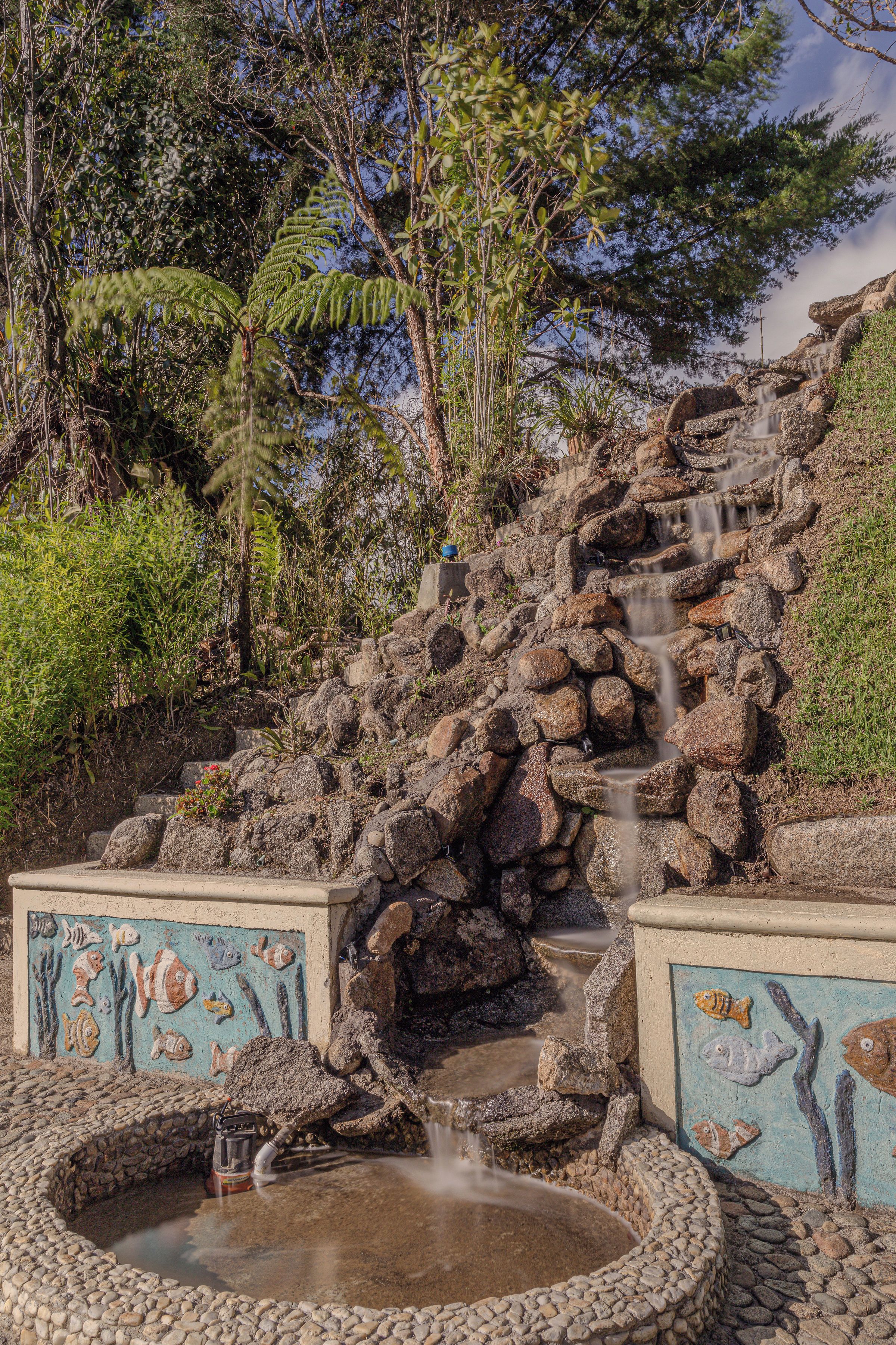 Una cascada natural de roca que desemboca en una piscina circular, rodeada de vegetación exuberante y una pared pintada con motivos de peces.