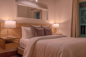A hotel room with a bed, two bedside lamps, a nightstand, a wooden headboard, and a mirror above the bed.