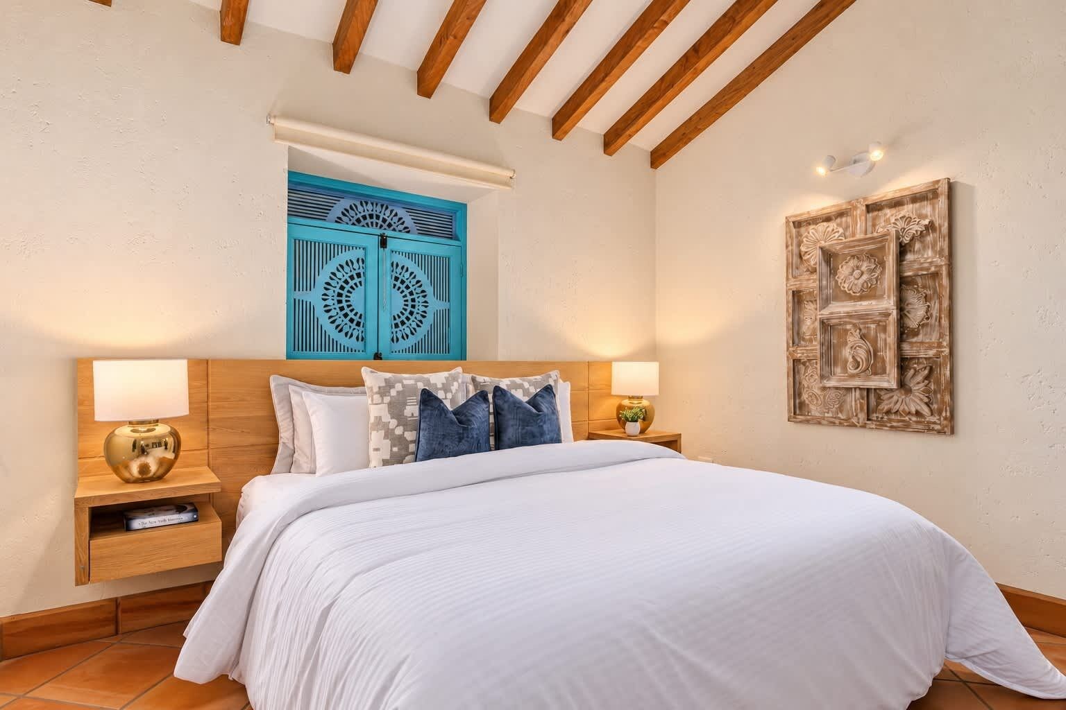 A bedroom with a large bed, two bedside tables with lamps, a wooden ceiling, and a decorative wall piece.