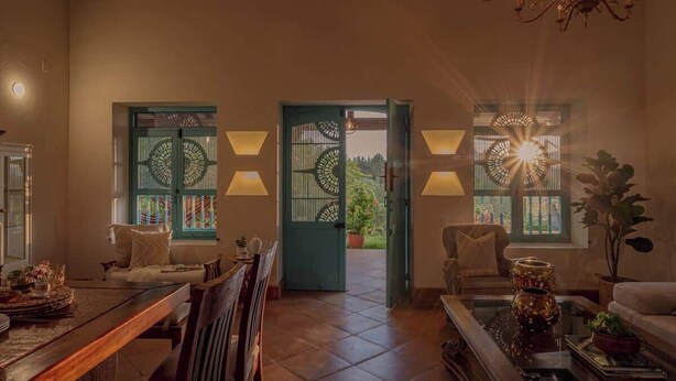 Uma sala de estar aconchegante com área de jantar, um sofá, uma mesa de centro e um candelabro. Há duas janelas com portões decorativos e uma porta que dá para o exterior.