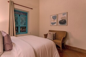 A bedroom with a bed, a chair, a window with blue shutters, and two wall-mounted artworks.