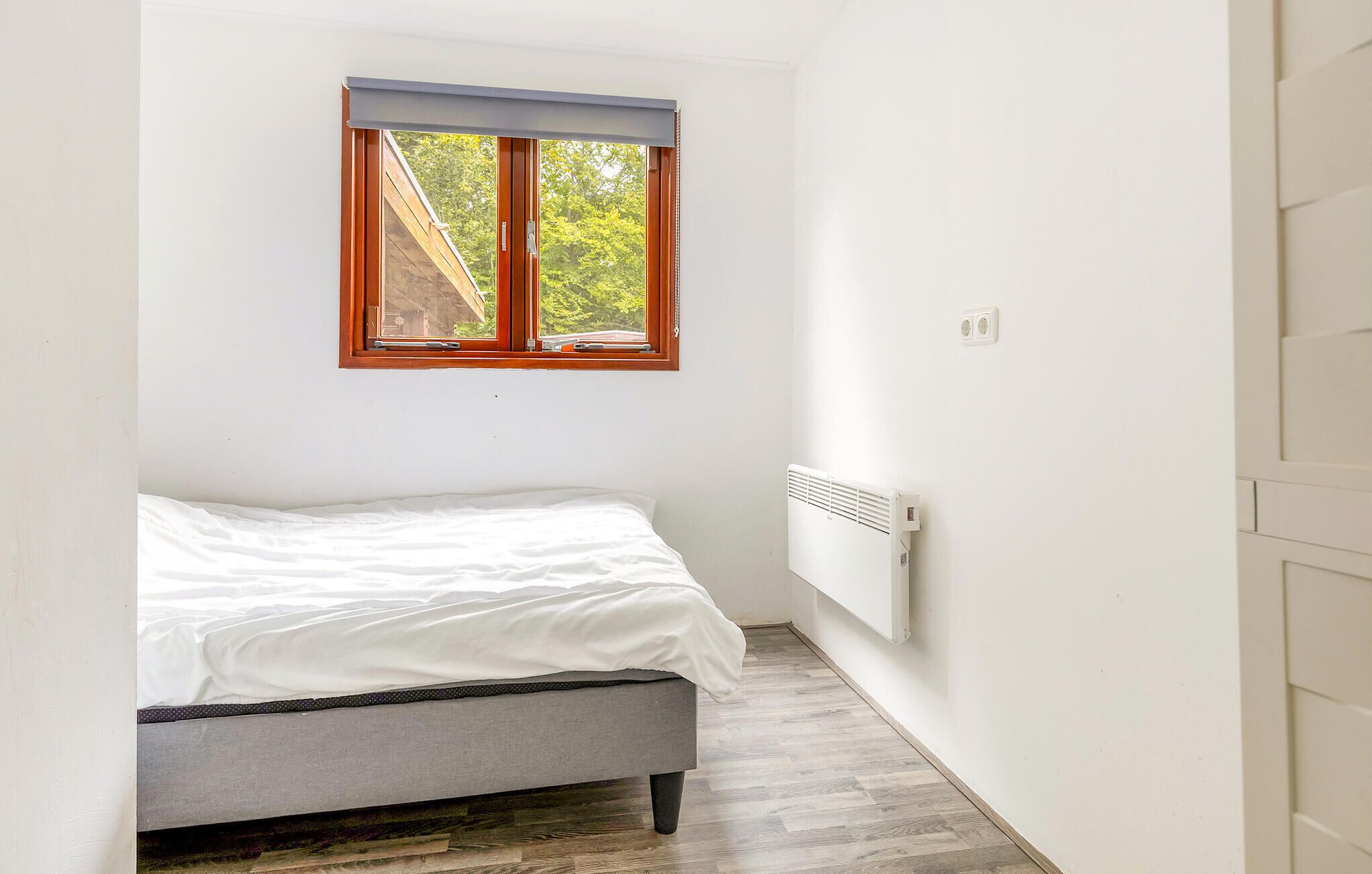 Une petite pièce lumineuse, avec un lit simple, une fenêtre donnant sur des arbres et un radiateur au mur.