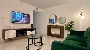 A modern living room with a green sofa, a glass coffee table, a TV mounted on the wall, a wooden cabinet, and a floor lamp. - Beautiful 2/2.5 Condo w/free parking fast WiFi (Miami)