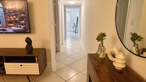 A hallway with a tiled floor, a wooden console table with decorative vases, a large mirror, and a flat-screen TV displaying a bookshelf scene. - Beautiful 2/2.5 Condo w/free parking fast WiFi (Miami)
