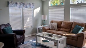 A living room with a brown leather sofa, a white coffee table, and a dark brown armchair. There is a ceiling fan, a window with blinds, and a small table with a lamp.