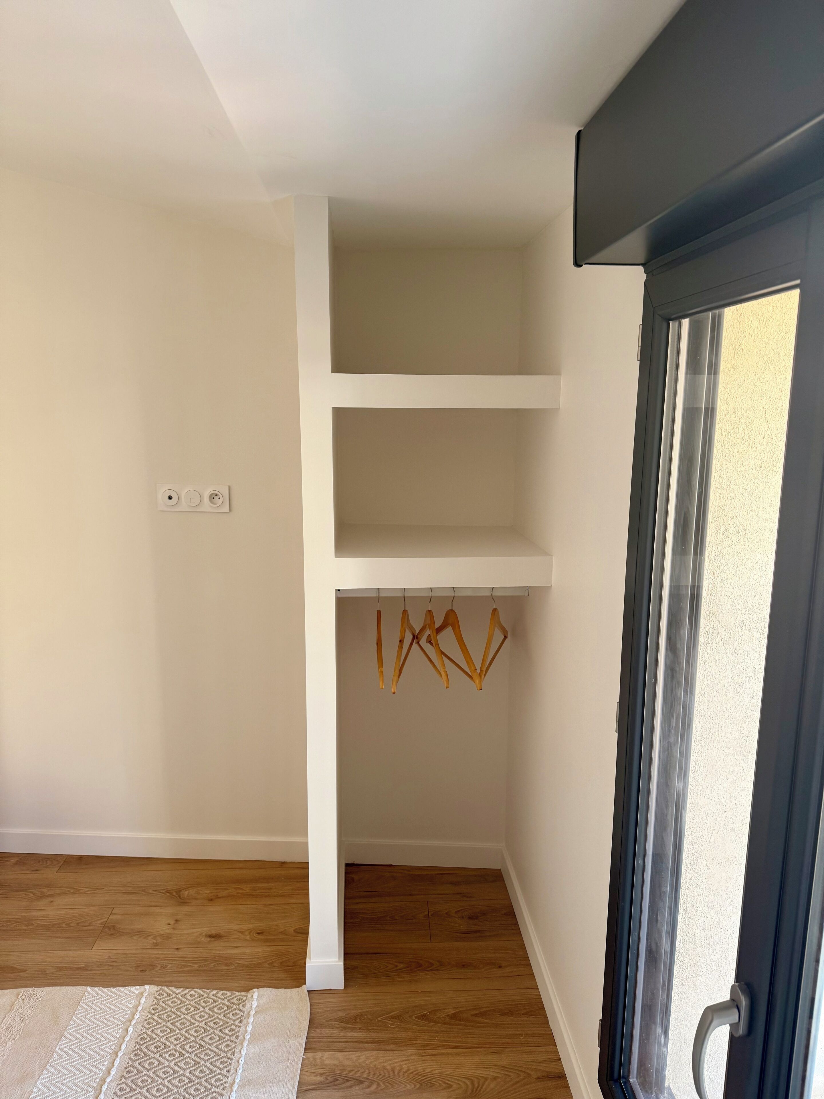 Une pièce avec un plancher en bois, des murs blancs et une porte dotée d’un panneau en verre. On y trouve trois étagères blanches et un ensemble de cintres en bois fixés sur une tringle.