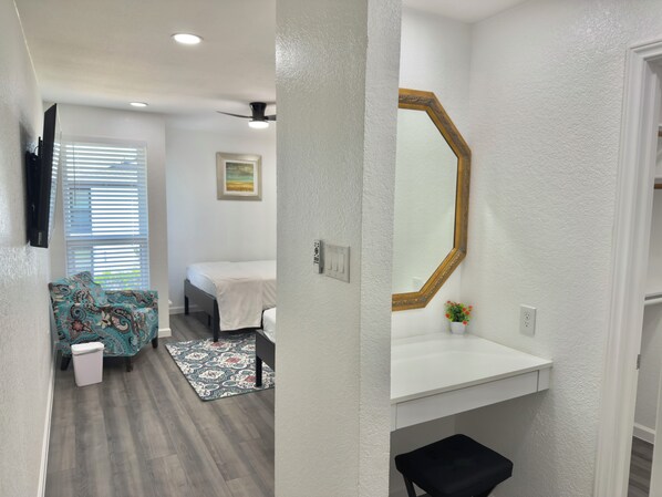 A hotel room with a bed, a bathroom with a sink and mirror, a small stool, and a window with blinds.