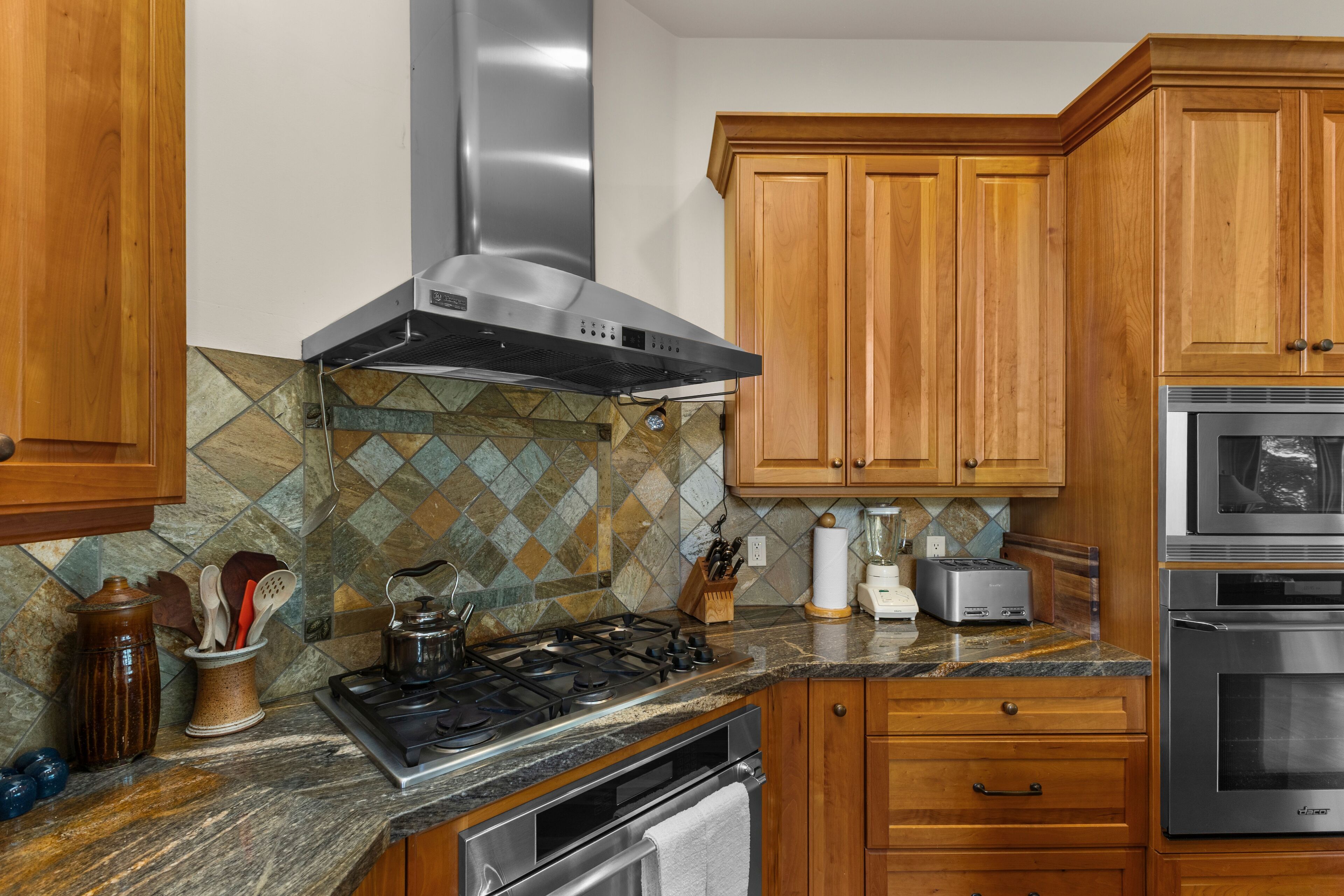 A kitchen with wooden cabinets, a gas stove, a stainless steel range hood, a tiled backsplash, and modern appliances.